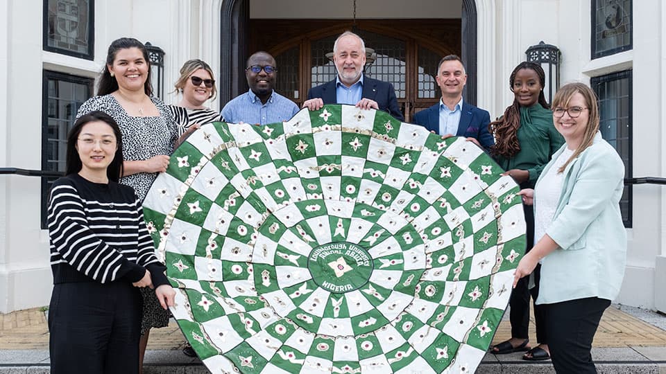 麻豆視頻_麻豆直播_麻豆传媒官网 staff from the Vice-Chancellor's Office, Alumni Relations Team and Global Engagement Team holding a tapestry made by alumni in Abuja  in front of Hazlerigg Building on 麻豆視頻_麻豆直播_麻豆传媒官网 campus. They are all smiling in the sunshine and standing on the steps outside the doorway.